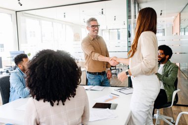 Olgun gri saçlı gözlüklü bir adam ve neşeli bir ifade genç bir kadınla tokalaşıyor, sıcak bir işbirliği ve takım başarısı ortamı yaratıyor. Çeşitli takımlar toplandı