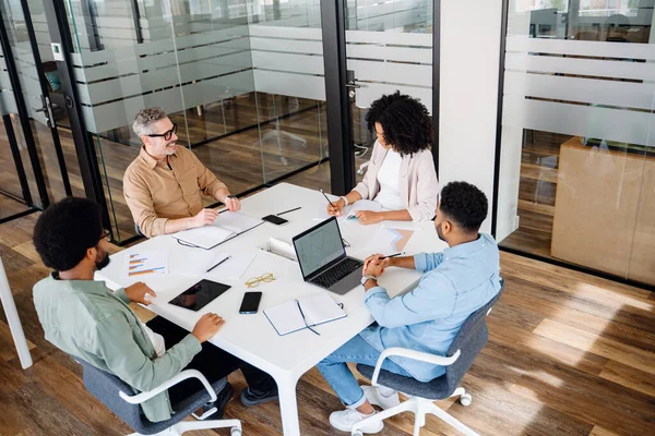 A team of four professionals are gathered around a white table in a modern office, indicative of a cooperative and strategic business meeting. Productive meetings and brainstorming concept