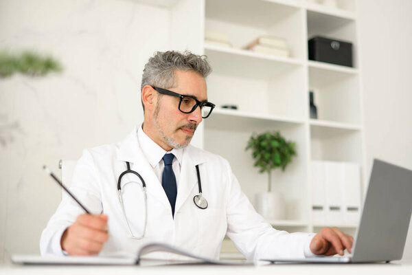Focused senior doctor with grey hair reviews patient information on a laptop and taking notes, symbolizing the integration of technology in modern medical practice, within contemporary clinic setting