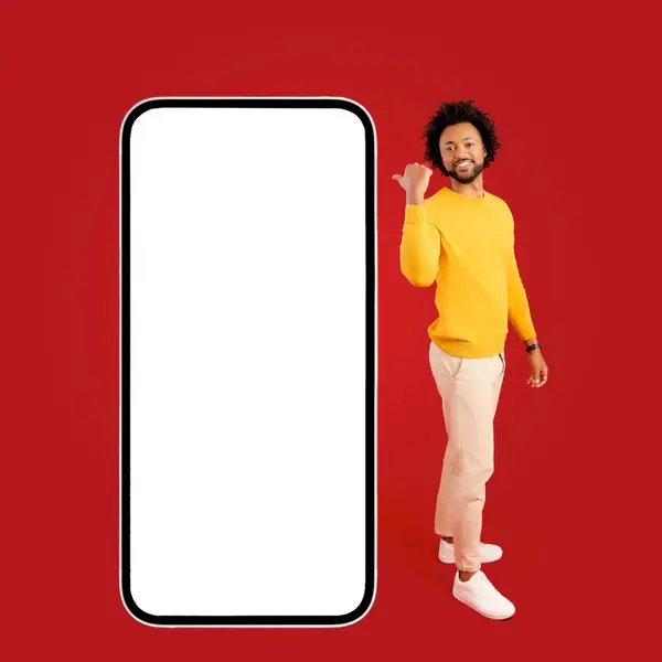 Look here. Handsome African-American curly man paying your attantion at huge mobile phone with empty screen isolated on red, charismatic guy point finger at smartphone with blank display, copy space