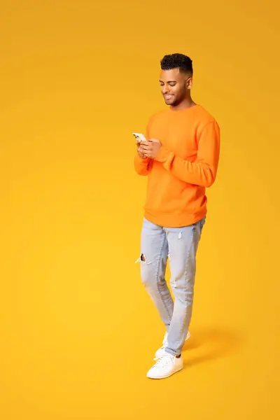 Full length photo of smiling young Indian man texting on the smartphone, using mobile phone for communication online, hindu male freelancer developer testing app on the phone isolated on yellow