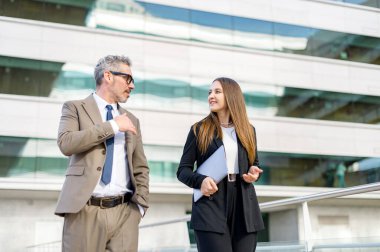 Tecrübeli iş adamı, bayan meslektaşı ile dışarıda konuşurken el kol hareketi yapar. İş kıyafetleri içinde kıdemli bir adam ve genç bir kadın arka planda modern ofis ile yürür ve sohbet ederler.