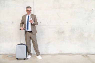 Akıllı telefonuyla meşgul olan işadamı, bavulunun yanında durarak, teknolojinin ve iş dünyasının bütünleşmesini sembolize ediyor. Seyahat ve iş konsepti