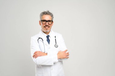 Dostça bir gülümseme ve profesyonel bir tavırla, bu kıdemli doktor, beyaz önlüklü, steteskoplu, temiz, beyaz bir arka plana karşı hazır bir şekilde yaklaşılabilirlik ve deneyim yayıyor..