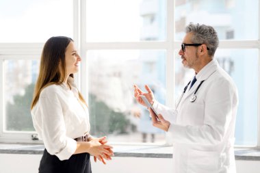 Üst düzey bir doktor, genç bir kadın hastayla sohbet ediyor. İkisi de güneş ışığında, modern bir muayenehanede tablete bakıyor. Bu resim hasta merkezli tedaviye olan bağlılığı yansıtıyor.