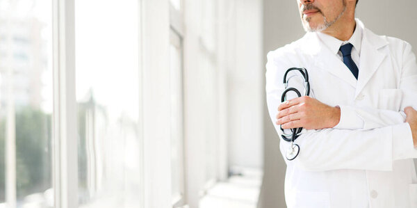 Задумчивый старший врач со стетоскопом, стоящий у окна в белом халате, его осанка передающая опыт и созерцание. Широкий баннер, обрезанный вид, лицо не видно
