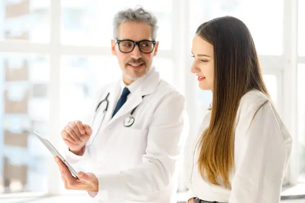 stock image A senior doctor, characterized by his grey hair and professional demeanor, uses a tablet to explain medical details to a receptive young female patient