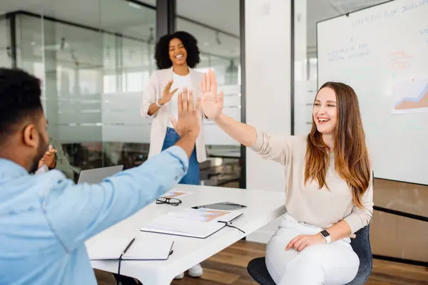 In a moment of triumph, a woman high-fives her male colleague, while another team member laughs in the background, capturing the essence of a goal achieved in a lively office brainstorming session