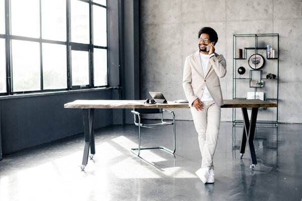 Modern Brazilian businessman speaks on the phone while casually leaning against a desk in a chic, sunlit office, exuding a vibe of casual professionalism. Concept of connectivity and business agility