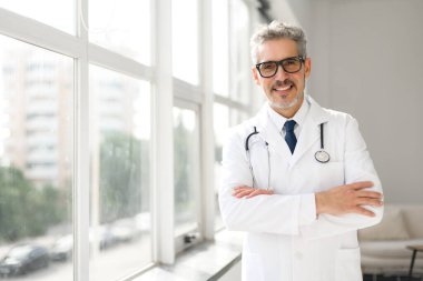 Dostça gülümseyen olgun bir doktor geniş ve parlak bir kliniğin arka planında durarak kollarını kavuşturur. Görüntü, güven ve yeterlilik hissi veriyor.