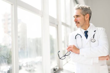 Beyaz önlüklü kıdemli bir doktor parlak bir klinik odasında duruyor, nazik bir ifadeyle ve gözlükleriyle, yoğun bir günün ortasında bir anlık dinlenmeyi ya da derin düşüncelere dalmayı yansıtıyor.