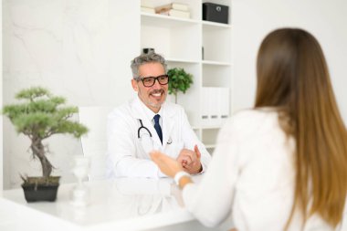 Parlak bir klinik ortamında, beyaz önlüklü kıdemli bir doktor bir hastayla konuşur, sağlık konseptlerini açıklamak için jestler kullanır, ilgi çekici ve bilgilendirici bir tıbbi konsültasyon gösterir.