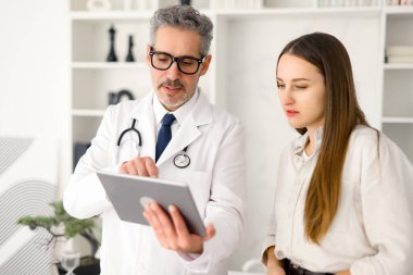 Yaşlı, gri saçlı bir erkek doktora, genç bir kadın hastaya dijital tablet kullanarak bilgi aktarırken gösteriliyor.