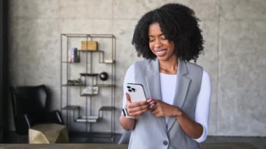 Tarz sahibi gri ceketli neşeli bir Afro-Amerikalı kadın modern ofis ortamında parlak bir gülümsemeyle akıllı telefonuna bakar, profesyonellik ve yakınlık yayar..