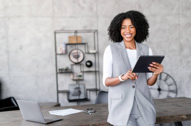 Özgüvenli bir Afro-Amerikalı iş kadını, iyi seçilmiş ofisinde, modern profesyonellik ve ulaşılabilirliğin bir karışımını vurgulayan parlak gülüşü ve günlük zarafetiyle bir tablet tutuyor.