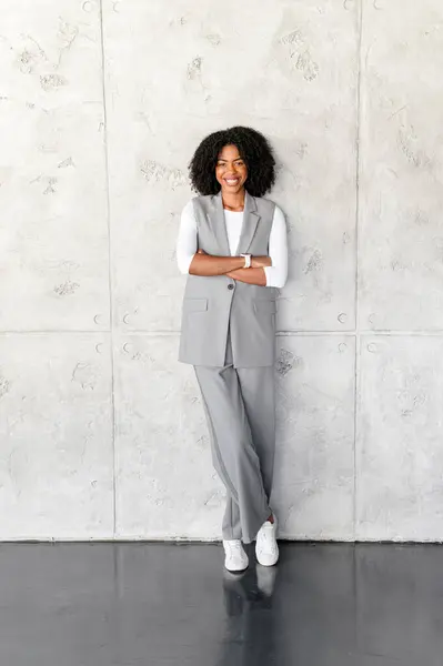 A professional African-American businesswoman stands confidently, arms crossed, with a grey wall on the background, exuding leadership and poise