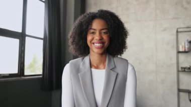 Işıl ışıl bir gülümseme ve dostça bir el sallamayla, Afro-Amerikalı bir kadın video görüşmesi sırasında meslektaşlarını selamlıyor. Modern, iyi aydınlatılmış ofis sanal iş toplantıları için ideal bir zemin sağlıyor.