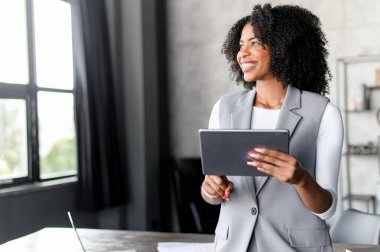 Geniş bir gülümsemeyle, Afro-Amerikalı bir iş kadını dijital tabletiyle çağdaş bir ofisin arka planında etkileşime giriyor. Şirket ortamında dijital bağlantının kolaylığı