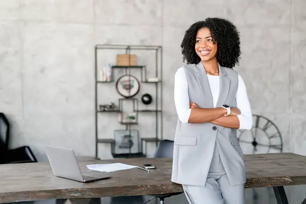 一位快乐的非洲裔美国女商人靠在办公室的桌子上 她的服装和举止彰显了专业精神和现代优雅 这个概念的重点是商业与风格的协调 — 图库照片