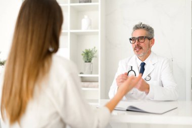 Olgun, gri saçlı, gözlüklü bir doktor bir hastayı dikkatle dinliyor ve ofisinin el değmemiş ortamında sağlık sektöründe iletişimin önemini vurguluyor..