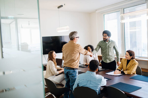 Команда кружит вокруг стола заседаний, где рукопожатие между двумя членами занимает центральное место, отражая момент согласия или достижения в рамках разнообразной и совместной рабочей группы