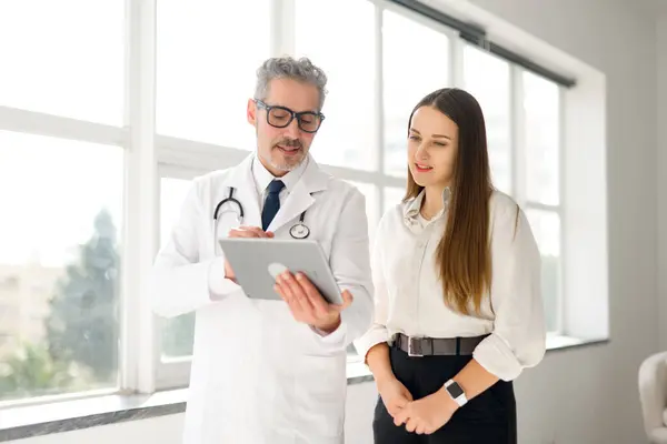 In this image, a mature doctor with a digital tablet discusses healthcare options with a young female patient in a well-lit room, illustrating the dynamic of modern medical consultation