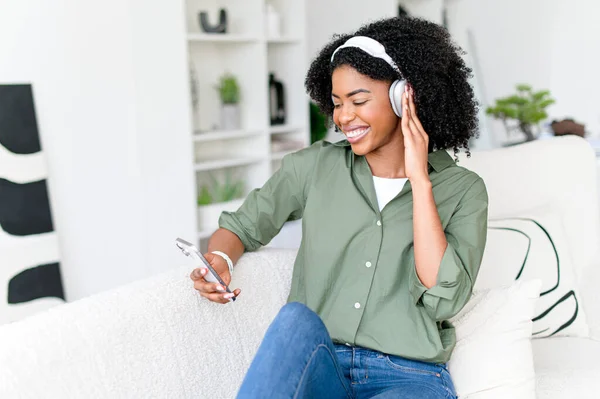 stock image Joyful African-American woman with curly hair enjoys listening to music on her headphones and smartphone while sitting on a couch. The role of technology in contemporary relaxation and entertainment.