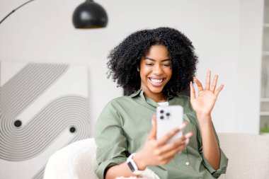 Neşeli bir dalga ve büyüleyici bir gülümsemeyle Afro-Amerikalı kadın, akıllı telefonuyla yaptığı bir video konuşmasında, modern, minimalist evinin rahatlığıyla çevrelenmiş bir şekilde birini selamlıyor gibi görünüyor.