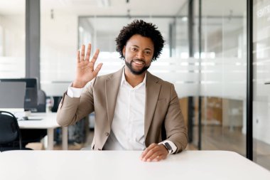 Parlak bir ofiste, yaklaşılabilir bir Afro-Amerikalı işadamı, iş arkadaşları veya iş arkadaşlarıyla sanal buluşmaya katılan dostane bir el hareketi yapar.