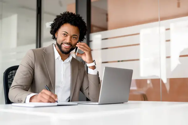 アフリカ系アメリカ人のビジネスマンは 彼が電話会話に従事するときの快適さとコントロールを排除し 彼の前にノートパソコンが開いているのに気をつけて座ります マルチタスクとリモート通信 — ストック写真