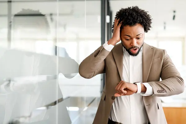 アフリカ系アメリカ人の実業家は 手首の時計をチェックし 企業生活の速い性質を反映した現代のオフィスに立っていることに不安を感じているようだ 緊急性あるいは遅刻するという概念 — ストック写真