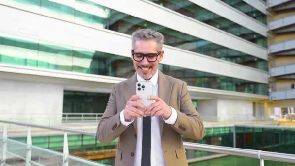 一位头发灰白的高管喜欢在他的智能手机上带着微笑在线发短信 在现代建筑的背景下阅读一份积极的业务更新报告 — 图库视频影像