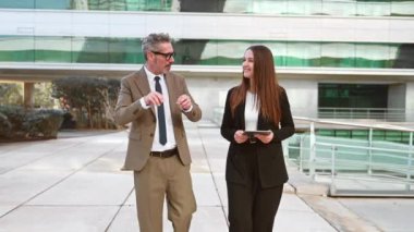 Gri saçlı bir işletme yöneticisi ve genç bir bayan ortak bir iş bölgesinde yan yana yürüyorlar, derin bir sohbete dalmışlar, aktif iş hareketliliği ve akıl hocalığı sergiliyorlar.