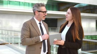 Deneyimli bir iş adamı, genç bir kadın profesyonel tarafından tutulan tablet ekranına bakarken gri saçlı gülümsüyor. Bu, kurumsal ortamdaki deneyimli uzmanlık ve modern teknolojinin birleşmesini temsil ediyor.