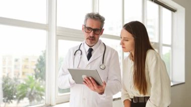 Beyaz önlüklü, gri saçlı bir doktor, parlak, modern bir sağlık hizmetleri yaklaşımını somutlaştıran, genç bir kadınla tablet hakkında bilgi paylaşıyor.