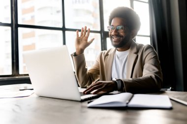 Genç bir ofis çalışanı, bilgisayardaki sanal toplantı sırasında internetten konuşan rahat ama profesyonel bir tavır sergiliyor. Hoş bir jest havası var. Bu da dostane ve verimli bir iş ortamı sağlıyor.