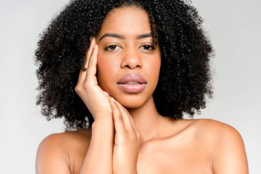 Afro-Amerikalı bir kadının yakından çekilmiş bir portresi, eli nazikçe yüzünü çerçeveliyor, tarafsız bir arkaplana karşı ayarlanmış düşünceli bir bakış ve yansıtıcı bir hava sergiliyor..