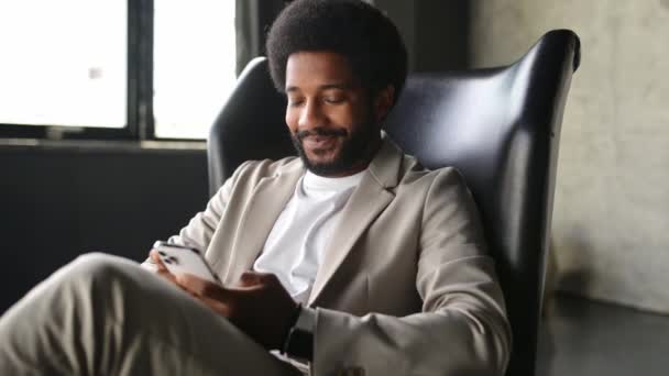 一位心情舒畅 面带微笑的巴西商人坐在时髦的办公椅上 一边在智能手机上享受着自己的时光 这象征着现代科技在商业领域的轻松发展 — 图库视频影像