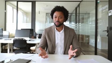 Parlak bir ofiste, yaklaşılabilir bir Afro-Amerikalı işadamı, iş arkadaşları veya iş arkadaşlarıyla sanal buluşmaya katılan dostane bir el hareketi yapar.