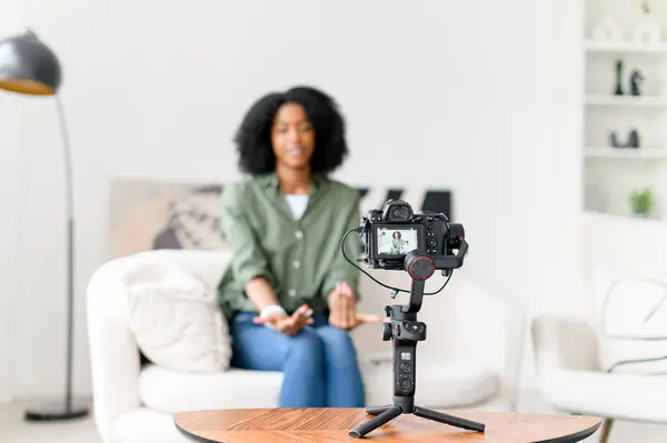 With a wave and a smile, an African-American woman engages her online audience, creating an inviting atmosphere in her vlog from a minimalist living space