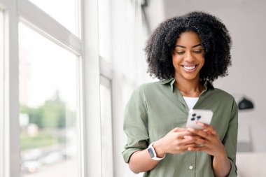 Işıl ışıl kapalı bir ortamda, bulaşıcı bir gülümsemesi olan Afro-Amerikalı bir kadın akıllı telefonuyla iletişim kuruyor, sosyal ağ üzerinden arkadaşlarıyla veya takipçileriyle iletişim kuruyor..