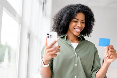 Akıllı telefon ve kredi kartı tutan Afro-Amerikalı bir kadın, muhtemelen online alışveriş veya rezervasyon yapıyor, parlak, modern bir iç mekanda.