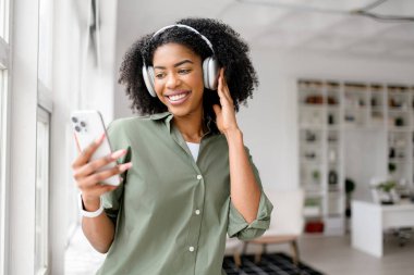 Rahat zeytin yeşili tişörtlü Afro-Amerikalı bir kadın beyaz kulaklıklarıyla müzik dinlerken ışıl ışıl gülümserken, modern, iyi aydınlatılmış bir iç mekânda akıllı bir telefon tutuyor..