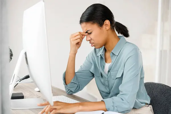 Exhausted mixed-race entrepreneur businesswoman or student closed her eyes touching massaging her eyes, suffering from severe headache migraine. Tired young woman overwhelmed with computer work