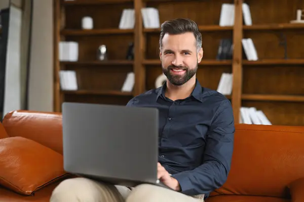 A freelancer man with a beard is sitting comfortably on the couch, using his laptop computer, looking at the camera with a smile on his face, types in a comfortable home office