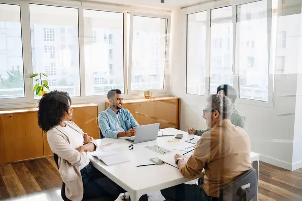 4人の専門家のチームが近代的なオフィスでホワイトテーブルの周りに集まり 協力的かつ戦略的なビジネスミーティングを示しています 多様なワークチームでガラスパーティションを見る — ストック写真