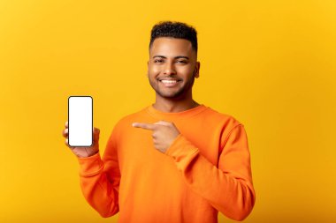 Surprised Indian man holding smartphone in hand and points finger at empty phone screen, astonishment guy presenting best deal, advertising new mobile app, isolated on yellow
