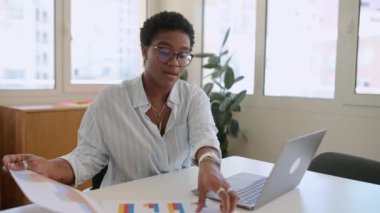 Neşeli genç bir profesyonel, güneşli bir ofis ortamında dizüstü bilgisayarından birini karşılıyor. Bu modern teknoloji ile dostça bir iş yeri iletişimi anı yakalar..