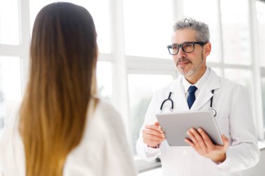 Gri saçlı olgun bir doktor, sağlık bilgilerini dijital tablette doğal ışıkla dolu bir klinikte genç bir kadınla tartışıyor.