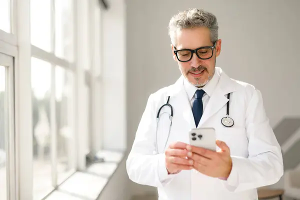 Dojrzały Siwowłosy Lekarz Bada Swój Smartfon Warunkach Klinicznych Prawdopodobnie Przeglądając — Zdjęcie stockowe